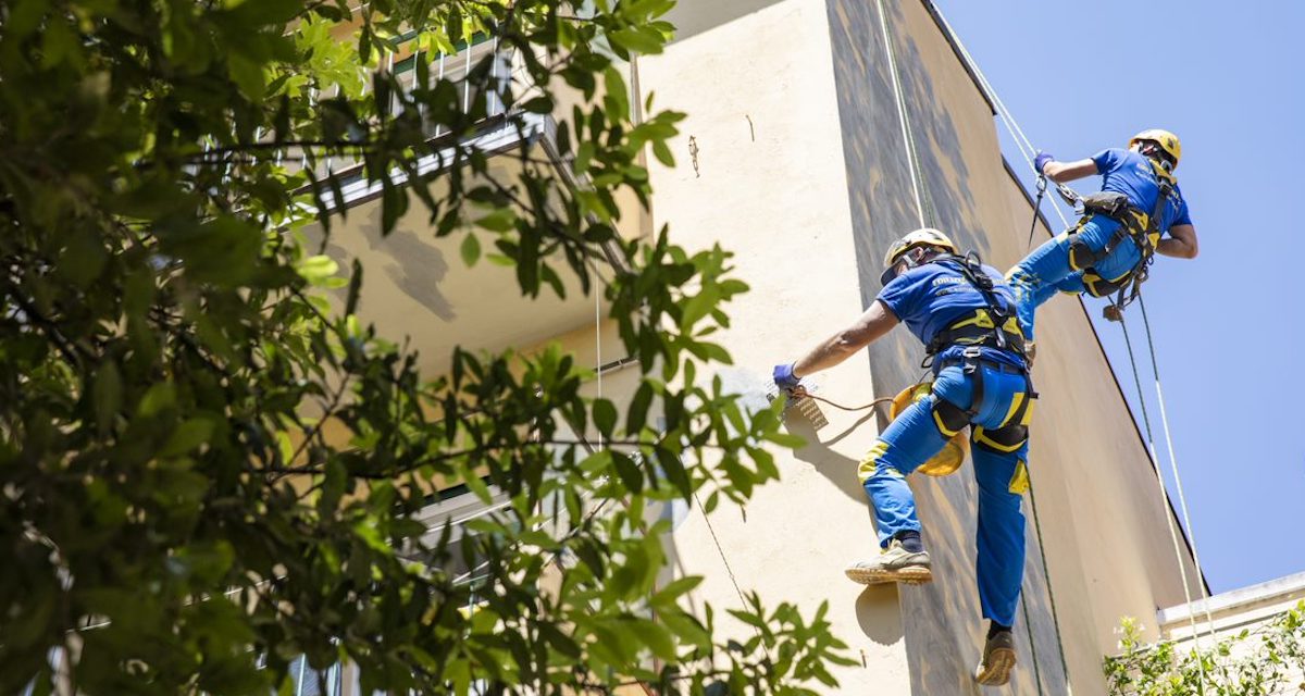 Foto di stock di Ediliziacrobatica. Due operai su fune che salgono verso il tetto di un'edificio per applicare potenzialmente la vernice al microcappotto termico